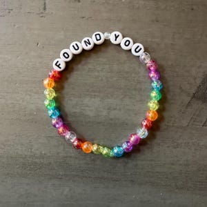 May include: A circular beaded bracelet featuring a sequence of translucent, multi-colored beads in shades of red, orange, yellow, green, blue, and purple. The top section includes white spherical beads with black letters spelling FOUND YOU.