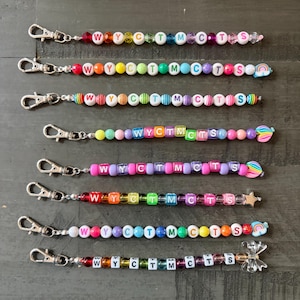 May include: Eight beaded lanyards featuring silver-toned lobster claw clasps. Each strand displays colorful plastic beads and letter beads spelling WYCTMCTS. The designs include various bead shapes, rainbow charms, a star, and a clear butterfly charm.