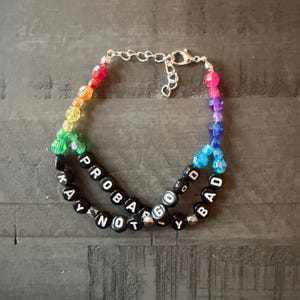 May include: A double-strand bracelet featuring a rainbow of faceted beads and black letter beads spelling out PROBABLY GOOD NOT BAD. The bracelet includes a silver-toned metal chain with a lobster claw clasp for secure closure and adjustment.