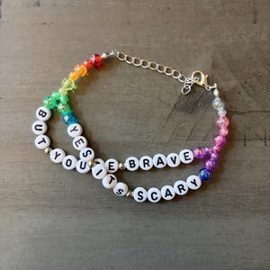 May include: A multi-strand bracelet featuring a rainbow of translucent beads and white letter beads spelling out the phrase YES BUT YOU IT'S BRAVE SCARY. The design includes a silver-toned metal chain with a lobster claw clasp and small metallic spacers.