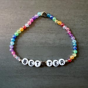 May include: A beaded bracelet featuring a circular arrangement of small, translucent, multi-colored beads. The bracelet includes white letter beads spelling out the word BETTER, with a silver-toned heart-shaped bead positioned between the letters.
