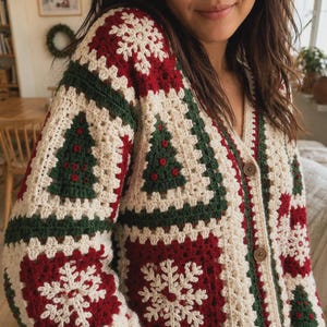 May include: A V-neck crochet cardigan featuring a patchwork design of cream, dark red, and forest green squares. The pattern includes white snowflake motifs and dark green Christmas tree shapes with red accents. The garment has two visible brown buttons.