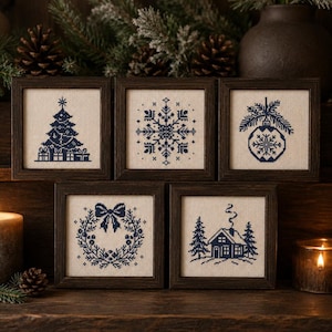 May include: Five square dark wood frames containing cross-stitch embroidery on cream fabric. The designs feature navy blue thread depicting a Christmas tree with gifts, a snowflake, a hanging ornament, a wreath with a bow, and a cabin in the woods.