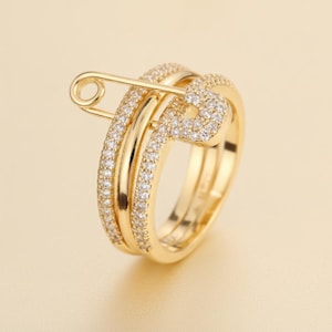 Peut inclure: Une bague dorée arborant un motif d'épingle à nourrice. L'anneau est incrusté de petites pierres claires et scintillantes sur les bords extérieurs, tandis que l'élément en forme d'épingle est également pavé de pierres assorties et brillantes.