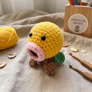 May include: A round, yellow crocheted creature with a pink circular mouth, two black bead eyes, and brown crocheted feet. A small green sphere is attached to the side. The figure is crafted from yarn and features a textured, knotted surface.