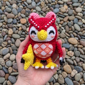 May include: A crocheted owl figure featuring a bright red body, a yellow beak, and yellow feet. The owl has pink ear tufts, a white face with black embroidered eyes, and a light pink and white diamond-patterned belly. It holds a yellow star.