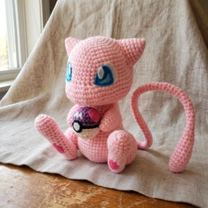 Puede incluir: Un peluche tejido a ganchillo de color rosa claro que representa una criatura felina con grandes ojos azules, orejas puntiagudas y una cola larga y delgada. La figura sostiene una pequeña bola redonda morada y blanca con un centro negro y una M blanca.