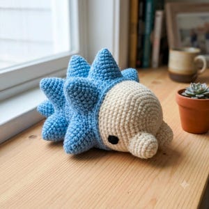 May include: A handmade crochet plush toy shaped like a hedgehog with a light blue spiky back and a cream-coloured face. The creature features a small, rounded snout and a single visible black bead eye. The texture is consistent with yarn stitching.