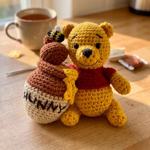 May include: A yellow crocheted bear wearing a red shirt, positioned next to a brown and cream crocheted honey pot with the word HUNNY embroidered on the front. A small crocheted bee sits on top of the honey pot, which features yellow yarn drips.
