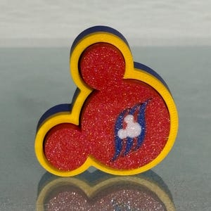 May include: A three-dimensional Mickey Mouse head icon featuring a bright yellow border and a red glitter-filled center. The center includes a blue wave-like graphic with a small white Mickey Mouse head silhouette integrated into the design.