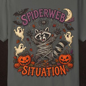 May include: A charcoal grey t-shirt featuring a graphic print of a raccoon tangled in a spiderweb. The design includes the text "SPIDERWEB SITUATION" in orange, surrounded by ghosts, bats, a crescent moon, and orange jack-o'-lantern pumpkins.