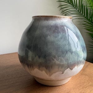 May include: A round ceramic vase with a wide body and a narrow opening. The surface features a glossy glaze with a gradient of deep blue and grey tones at the top, transitioning into a lighter, creamy off-white base with visible drip patterns.