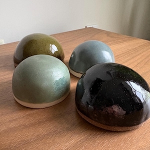 May include: Four ceramic half-sphere bowls with smooth, glazed surfaces in distinct colours: olive green, sage green, slate blue, and glossy black. Each bowl features a rounded dome shape with a flat base, showcasing a visible contrast between the coloured glaze and the exposed clay rim at the bottom edge.
