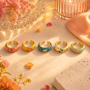 Peut inclure: Cinq bagues en métal doré disposées en rangée, chacune présentant un motif floral en émail unique. Les bagues affichent des couleurs vives dont le rose, le jaune, le bleu, la crème et le vert clair, avec des détails de fleurs et de feuilles peints.