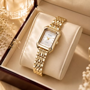 Könnte beinhalten: Eine goldfarbene analoge Armbanduhr mit rechteckigem Gehäuse und passendem Metallgliederarmband. Das Zifferblatt ist weiß mit goldfarbenen Zeigern und Indizes und zeigt den Markennamen RAGA sowie das Wort TITAN in schwarzer Schrift.