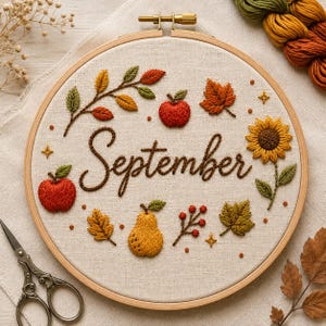 Puede incluir: Un bastidor de bordado con tela color crema que muestra la palabra September bordada en cursiva marrón. El diseño presenta elementos otoñales bordados, incluyendo manzanas rojas, una pera amarilla, un girasol, varias hojas y pequeñas bayas rojas.