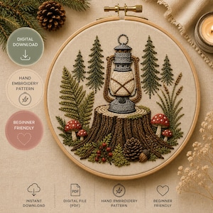 May include: A round wooden embroidery hoop containing a fabric panel with a hand-stitched design of a lantern resting on a tree stump. The scene includes embroidered pine trees, ferns, red-capped mushrooms, a pinecone, and small red berries.