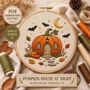 May include: An embroidery hoop containing a stitched design of a pumpkin house at night. The scene features a large orange pumpkin with a door and windows, surrounded by bats, a crescent moon, and fallen leaves on a cream fabric background.