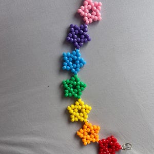 Può includere: Una catena di perline con sette ciondoli a forma di stella disposti in sequenza verticale. Ogni stella è composta da piccole perline di plastica di un colore distinto, seguendo uno spettro arcobaleno di rosa, viola, blu, verde, giallo, arancione e rosso.