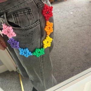 May include: A chain accessory made of multi-coloured plastic beads arranged in star shapes, featuring a rainbow colour sequence of red, orange, yellow, green, blue, purple, and pink, attached to the belt loops of grey denim jeans with metal clips.