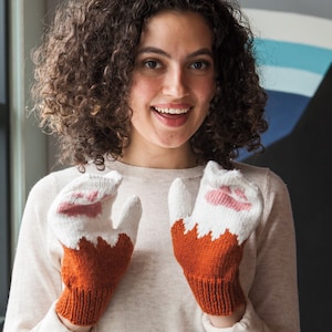 May include: A person wearing a white sweater and a pair of orange and white knitted mittens with pink paw prints. The mittens are shaped like cat paws.