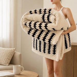 Puede incluir: Una manta de felpa color crema con un patrón geométrico negro repetitivo de rayas verticales en forma de chevrón. La tela tiene una apariencia gruesa, texturizada y similar al vellón, doblada en una forma rectangular grande y suave.