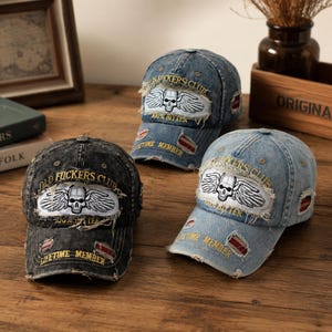 Puede incluir: Tres gorras de béisbol de mezclilla desgastada en negro, azul medio y azul claro. Cada gorra presenta un parche bordado central con una calavera alada, el texto OLD FUCKERS CLUB, 100% BITTER y LIFETIME MEMBER en hilo amarillo.