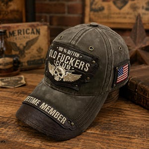 Puede incluir: Una gorra de béisbol gris oscuro desgastada con un parche marrón oscuro en la parte frontal con el texto 100% DITTER OLD FUCKERS CLUB y una calavera con alas. La visera muestra el texto LIFETIME MEMBER y tiene una pequeña bandera de EE. UU. al lado.
