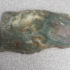 May include: A flat, rectangular slab of polished stone featuring a marbled pattern with blended shades of forest green, earthy brown, muted gray, and cream. The surface displays natural mineral inclusions and organic, swirling color variations.