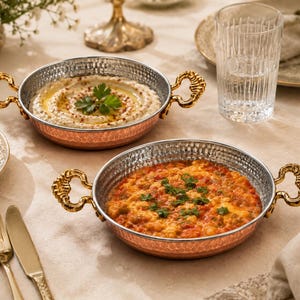 May include: Two round, shallow copper serving pans with hammered exterior textures and silver-toned interiors. Each pan features two ornate, gold-coloured metal handles on opposite sides. The pans contain prepared food garnished with fresh green herbs.