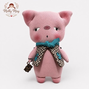 Könnte beinhalten: Eine rosa Stoffpuppe in Schweineform mit gestickten Augen, Wimpern und Schnauze. Sie trägt einen türkisfarbenen Schal und eine braun-weiß karierte Weste. Die Puppe hält einen kleinen, dunkelbraunen rechteckigen Anhänger mit Schleifendetail.