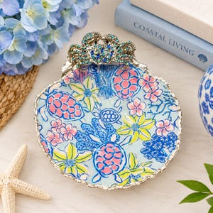 Può includere: Un piatto a forma di conchiglia con bordo argentato e un decoro a forma di granchio incastonato di cristalli blu e verdi. L'interno presenta un motivo colorato di tartarughe rosa, fiori gialli e corallo blu su uno sfondo azzurro.