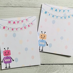 May include: Two rectangular birthday cards featuring a hand-drawn style robot and a banner that reads HAPPY BIRTHDAY. One card displays a pink and purple robot with pink banner letters, while the other shows an orange and blue robot with blue letters.