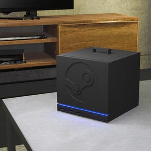 Może przedstawiać: Czarna, sześcienna konsola komputerowa z wytłoczonym logo Steam na przednim panelu. Urządzenie posiada mały uchwyt na górnej powierzchni oraz poziomy niebieski pasek świetlny LED umieszczony nad przednimi portami wejściowymi.
