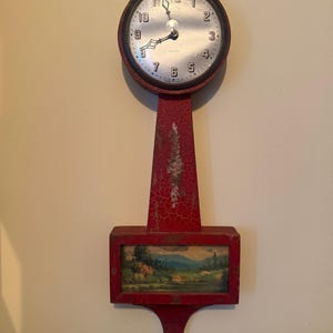 May include: A wall clock with a circular face featuring black numerals and hands, set within a red frame with a cracked paint finish. Below the clock face is a rectangular section displaying a landscape painting of trees and hills.