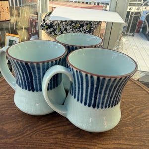 May include: Three ceramic mugs with a light blue base and a dark blue vertical striped pattern around the upper exterior. Each mug features a curved handle and a brown rim. The interior of the mugs is a solid light blue color, matching the base.