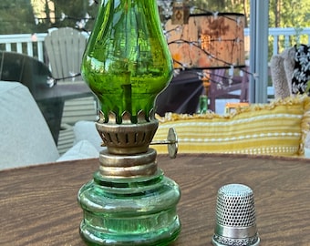 May include: A miniature oil lamp with a vibrant green glass chimney and base, featuring a metallic burner assembly with a side adjustment knob, positioned next to a small, silver-toned metal thimble with a textured, dimpled surface.