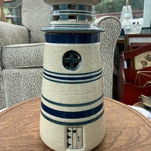 May include: A ceramic lighthouse sculpture featuring a cream-colored body with horizontal dark blue stripes. The structure includes a circular window with a cross design, a small rectangular door at the base, and a tiered top section with cutouts.