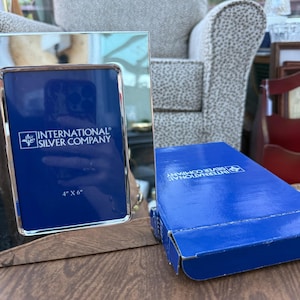 May include: A silver-toned metal picture frame with a reflective surface and a blue insert featuring the text International Silver Company and 4 x 6 inches. Beside the frame sits a matching blue cardboard box with the same brand logo.