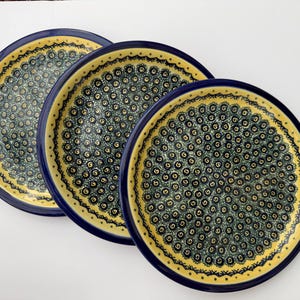 May include: Three round ceramic plates featuring a dark blue rim and a central pattern of small, repeating dark blue and green swirls on a cream-colored background. Each plate has a decorative border of small dots and lines along the inner edge.