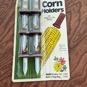 May include: A set of 6 corn holders with brown handles featuring gold flower accents and stainless steel prongs, packaged on a cardboard backing with an illustration of an ear of corn and the text Corn Holders with STAINLESS STEEL PRONGS.
