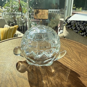 May include: A clear glass turtle figurine featuring a textured, geometric shell pattern. The object has a smooth, rounded head and a small tail, with a transparent body that catches the light, creating a distinct shadow on the surface below.