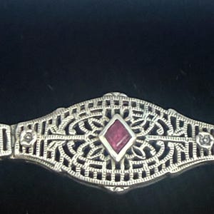 May include: A silver-toned bracelet link featuring an intricate openwork filigree design. The center of the piece contains a single, small, deep red diamond-shaped stone. Two tiny circular accents are positioned on either side of the central stone.