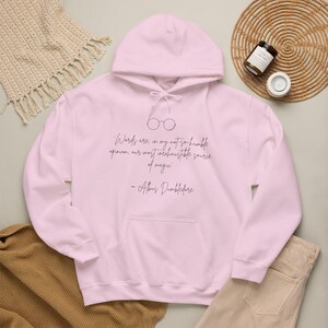 Può includere: Una felpa con cappuccio rosa chiaro caratterizzata da un disegno a linee nere di occhiali rotondi e la citazione: 'Words are, in my not-so-humble opinion, our most inexhaustible source of magic' seguita dalla firma '- Albus Dumbledore' in nero.
