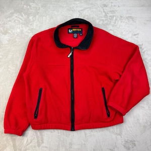 Pode incluir: Um casaco de fleece vermelho brilhante com gola alta preta, fecho de correr frontal preto a todo o comprimento e dois bolsos laterais verticais com fecho. A etiqueta interior da gola apresenta o texto EBTEK em letras brancas.