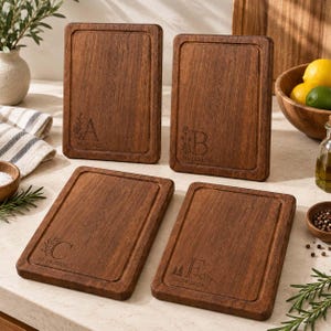 Puede incluir: Cuatro tablas de cortar rectangulares de madera marrón oscuro con esquinas redondeadas y un borde ranurado. Cada tabla presenta una letra grabada con láser, A, B, C o F, junto a un pequeño gráfico botánico o de montaña y la fecha 10.24.2026.