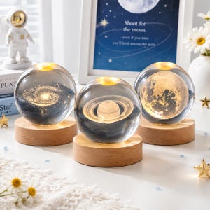 Op de afbeelding: Drie heldere glazen bollen rustend op ronde, lichtgekleurde houten voetstukken. Elke bol bevat een lasergegraveerde 3D-weergave van een hemellichaam: een spiraalstelsel, de planeet Saturnus met ringen en het textuurrijke oppervlak van de maan.