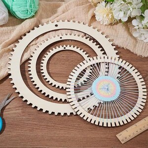 May include: Three circular wooden weaving looms with notched edges, arranged in descending size. The smallest loom is partially threaded with pastel-coloured yarn in a circular pattern. A metal pair of scissors and a wooden ruler are visible.