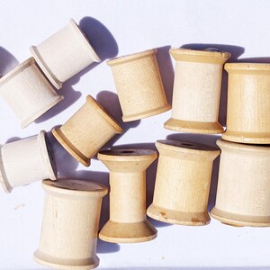 May include: A collection of wooden spools in various shades of beige and tan, scattered on a white surface. The spools are cylindrical with rounded edges, suggesting a craft or sewing theme.