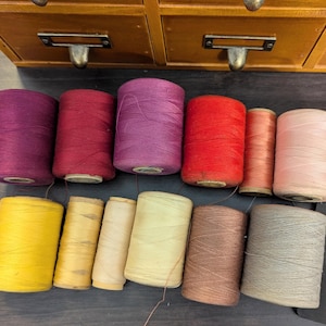 May include: A collection of 11 spools of thread in various colors, including red, pink, purple, yellow, and beige. The spools are arranged in two rows on a dark surface.